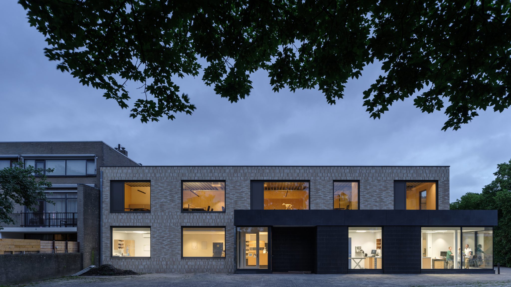Evening side front view corner building with glass corner aluminum window frames on ground floor extension. Light colored brick on CLT wood wall and new main entrance to veterinary clinic. Veterinary clinic design for Anicure ‘t Leidse Land in Leiderdorp, designed and built by MOST Architecture, Rotterdam