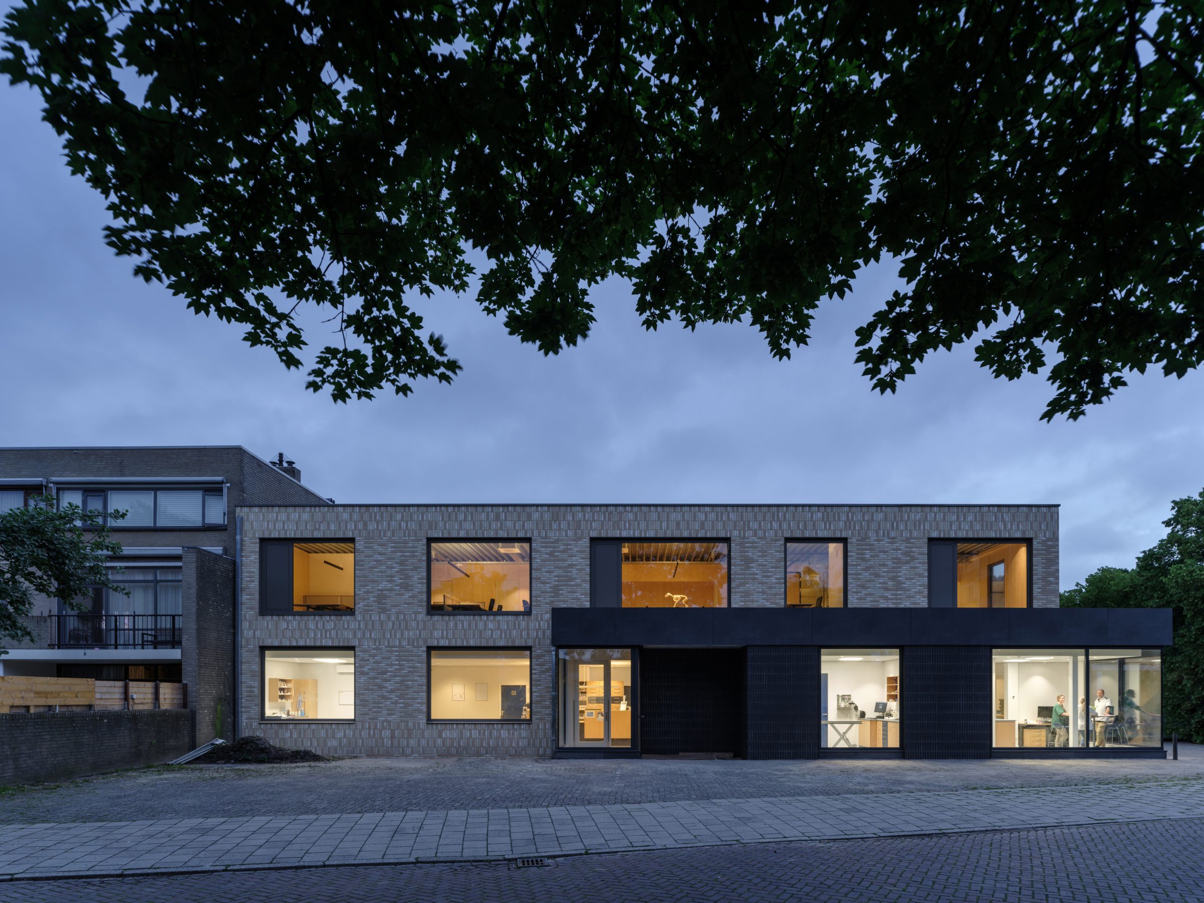 Evening side front view corner building with glass corner aluminum window frames on ground floor extension. Light colored brick on CLT wood wall and new main entrance to veterinary clinic. Veterinary clinic design for Anicure ‘t Leidse Land in Leiderdorp, designed and built by MOST Architecture, Rotterdam